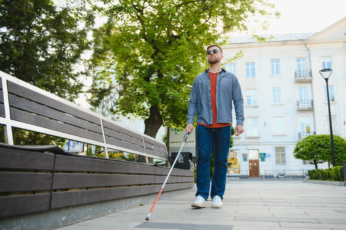 Photo d’un homme portant des lunettes de soleil qui marche sur un trottoir en ville avec une canne blanche longue, devant un banc de parc et des arbres