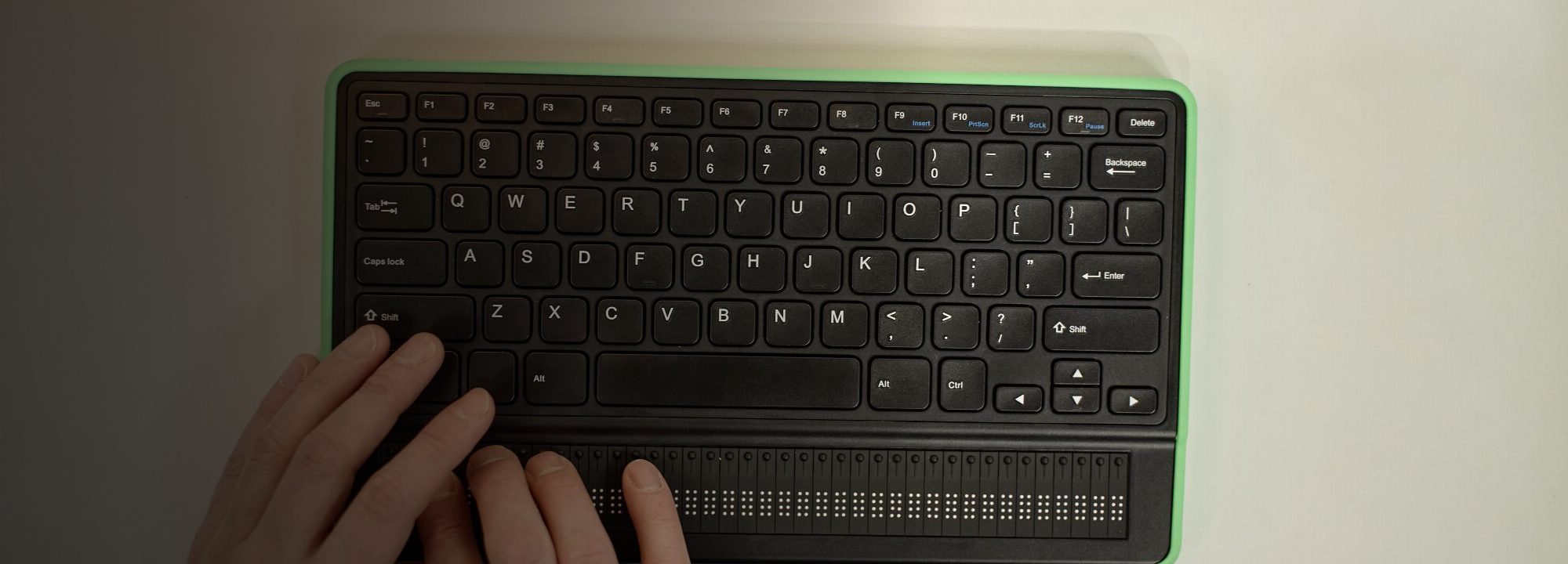 Vue de dessus : deux mains reposent sur un afficheur braille doté d’un clavier d’ordinateur intégré, le bout des doigts posé sur sa rangée de cellules braille.