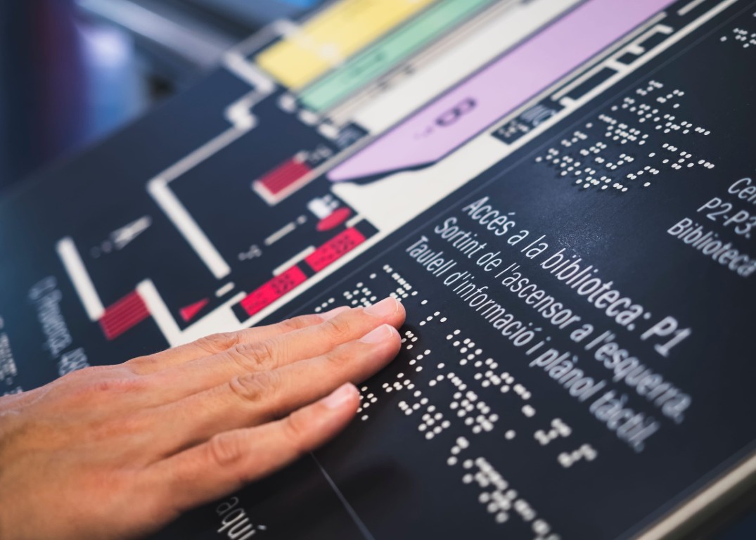 Photo d’une main qui lit le braille sur un plan d’orientation tactile, à côté d’un plan d’étage en relief et d’indications imprimées