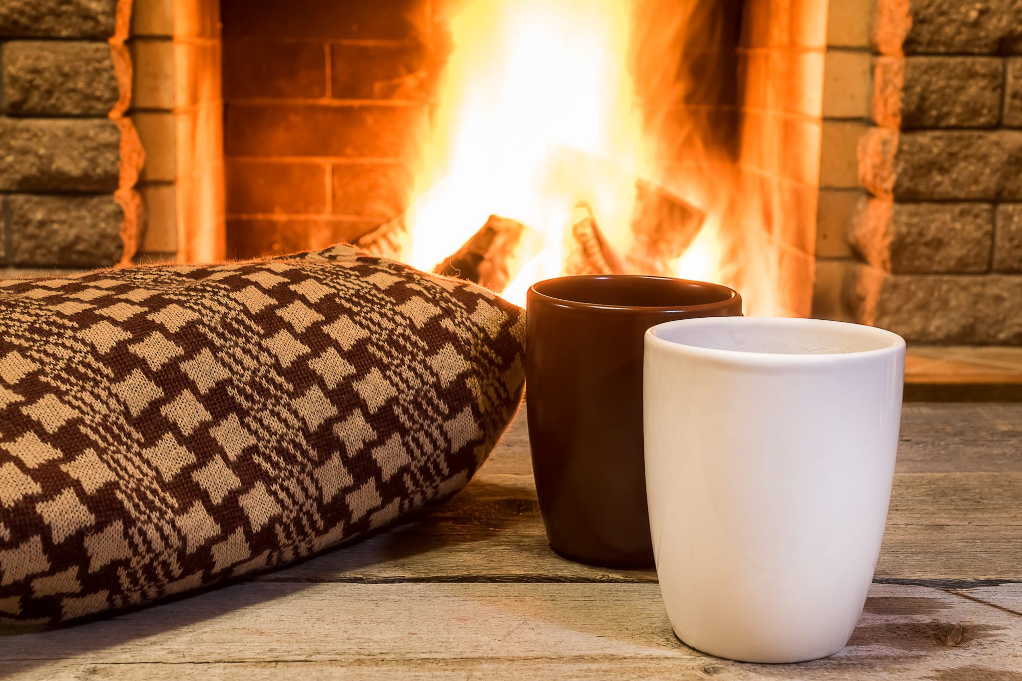 Photo d’une tasse brune et d’une tasse blanche posées sur un plancher de bois, à côté d’un coussin tricoté, devant un feu crépitant dans un foyer en pierre