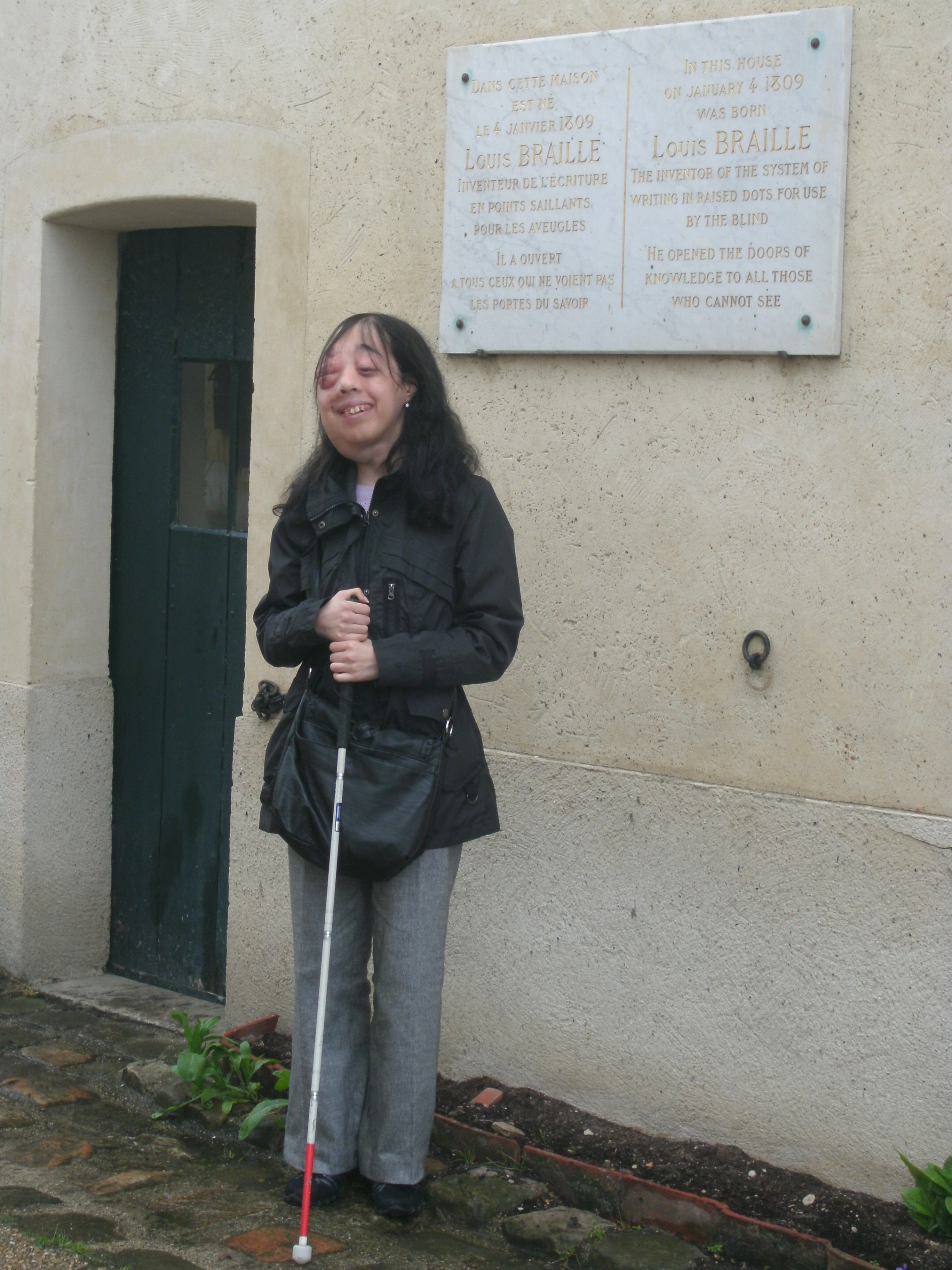 Natalina Martiniello se tient debout, souriante, sa canne blanche à la main, devant la maison en pierre de Coupvray, en France, où Louis Braille est né. Une plaque de marbre fixée au mur à côté d’elle indique, en français et en anglais, que Louis Braille — inventeur du système d’écriture en relief destiné aux personnes aveugles — est né dans cette maison le 4 janvier 1809.