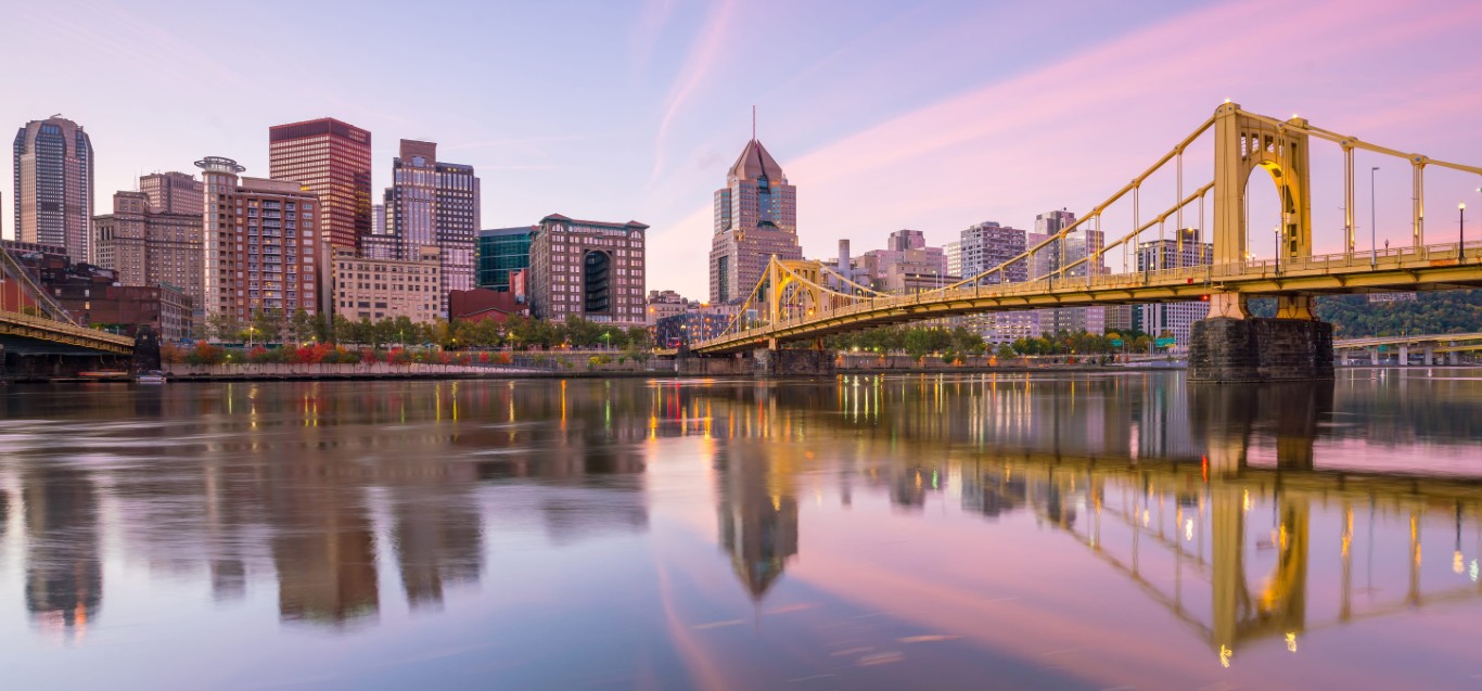 Photo de la silhouette urbaine de Pittsburgh au crépuscule, avec un pont jaune qui enjambe la rivière et se reflète dans l’eau rosée