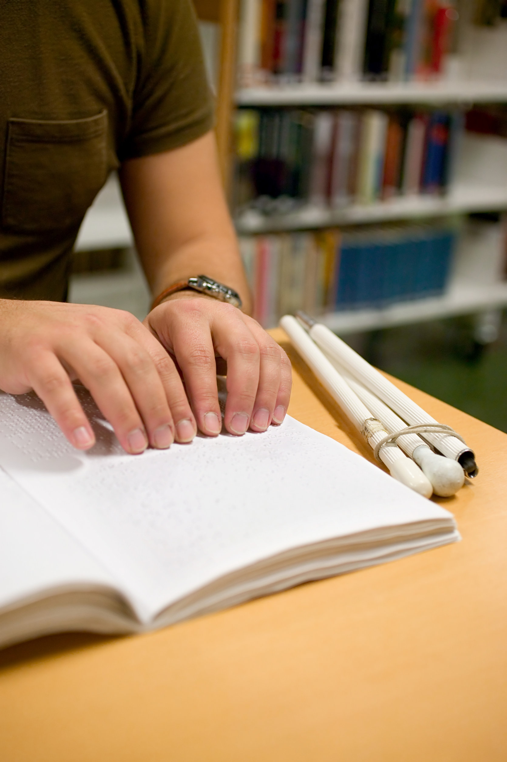 Photo d’une personne qui lit un livre en braille à une table de bibliothèque, une canne blanche pliée à côté du livre et des rayons de livres derrière elle